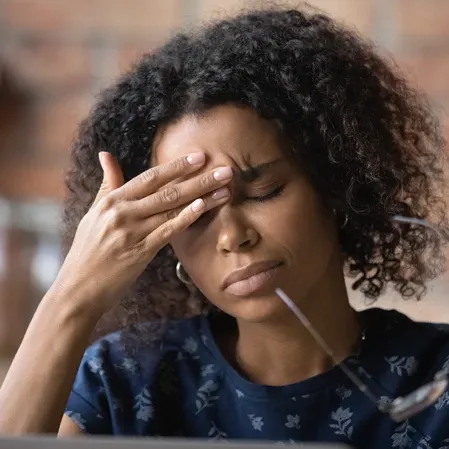 Person touching the forehead while seated, with eyeglasses visible in the foreground near a computer screen.