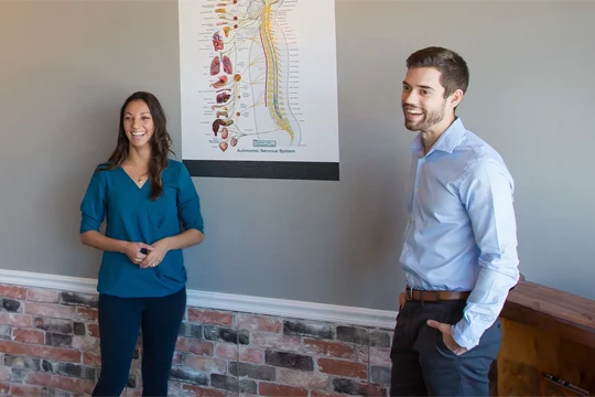 Dr. Tyler Leopardi and Dr. Brittany Leopardi standing near a wall with an anatomical poster displayed in the background.